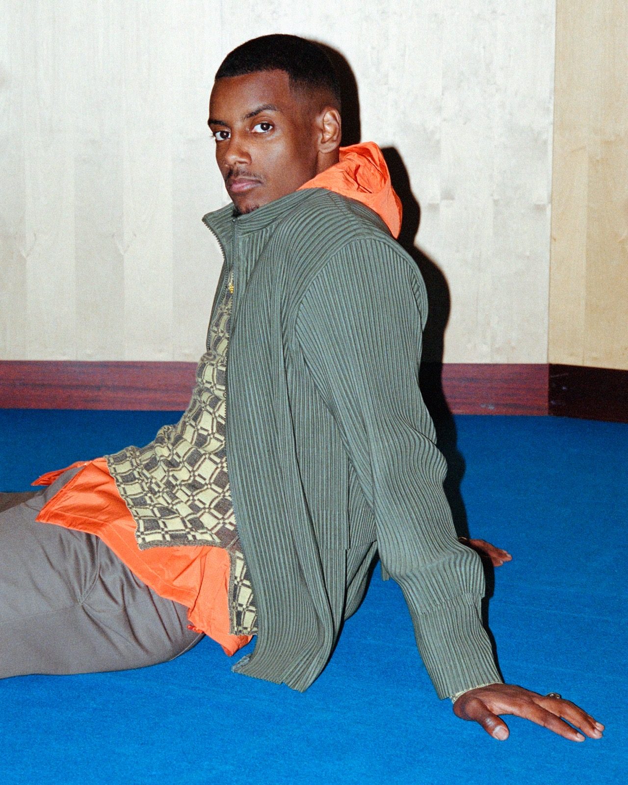 Alexander Isak seated on a blue floor in a ribbed cardigan and orange hoodie