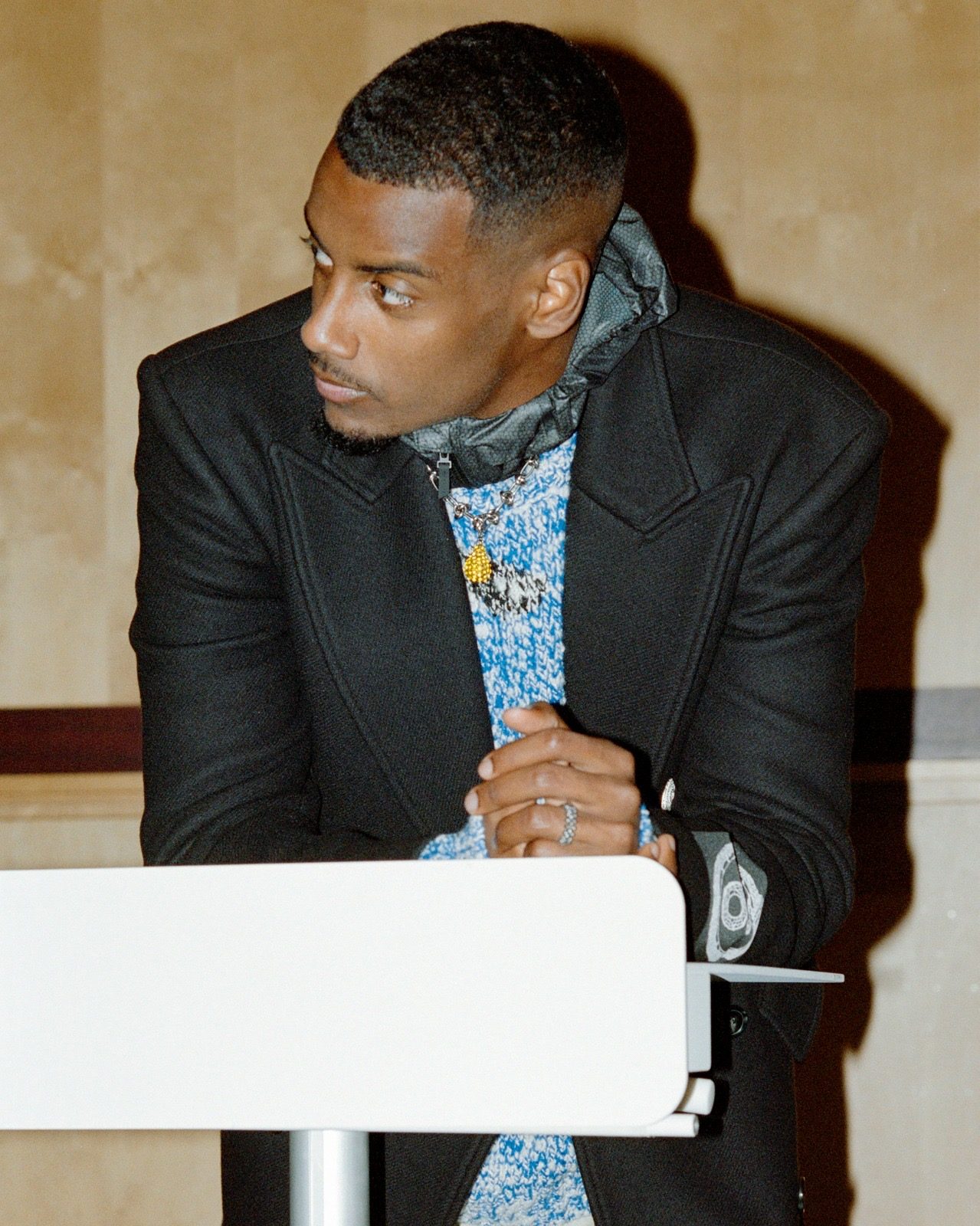 Alexander Isak in a black blazer and blue paisley shirt, leaning on a white chair in a warm interior